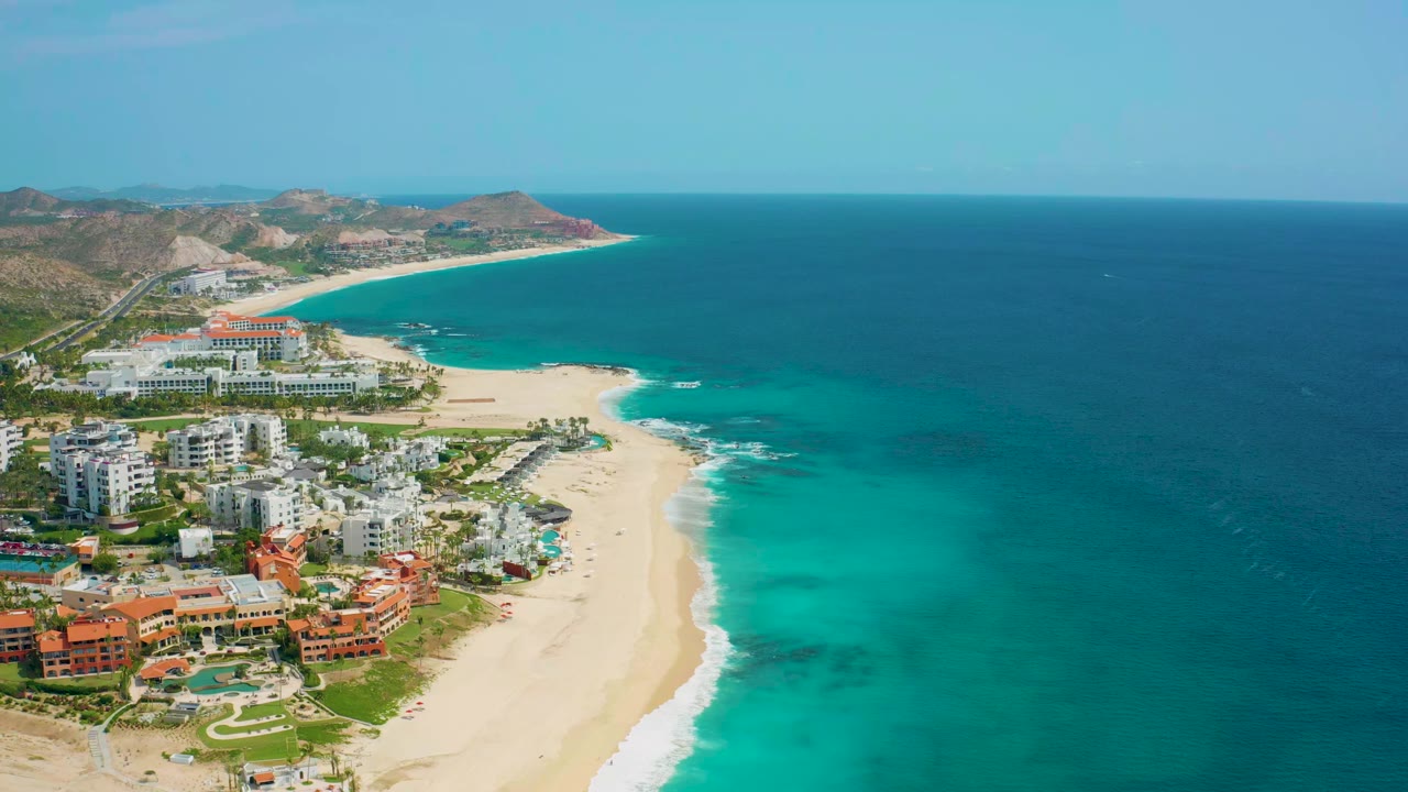 Opening aerial view over Desert Park Natural Reserve in Baja California Sur, Mexico, with resort buildings, a pale sandy beach, turquoise surf, arid hills, and no visible people.
