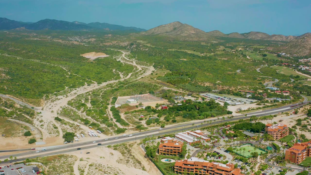 Opening aerial frame over Desert Park Natural Reserve in Baja California Sur, with a highway in the foreground, dirt tracks through green hills, a rocky peak beyond, and no visible people.