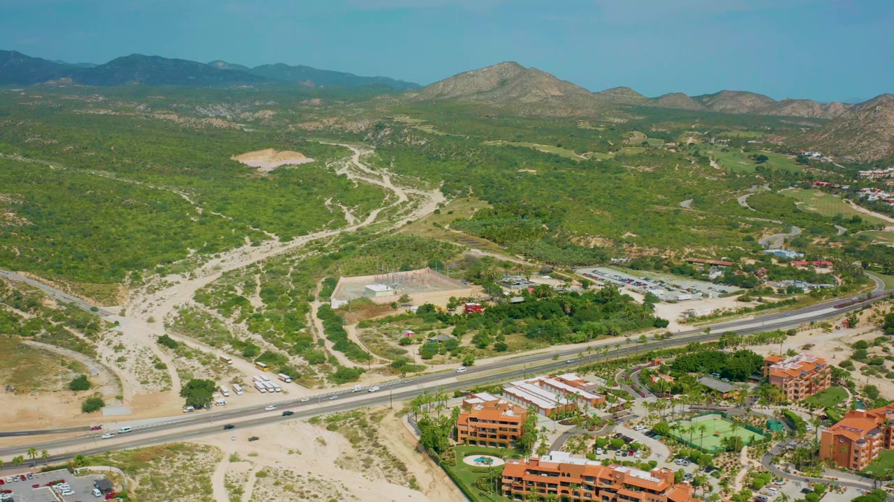 Aerial frame over the Baja California Sur reserve area showing the highway running across the lower frame, orange buildings near the road, pale dirt roads, green hills, and no visible people.