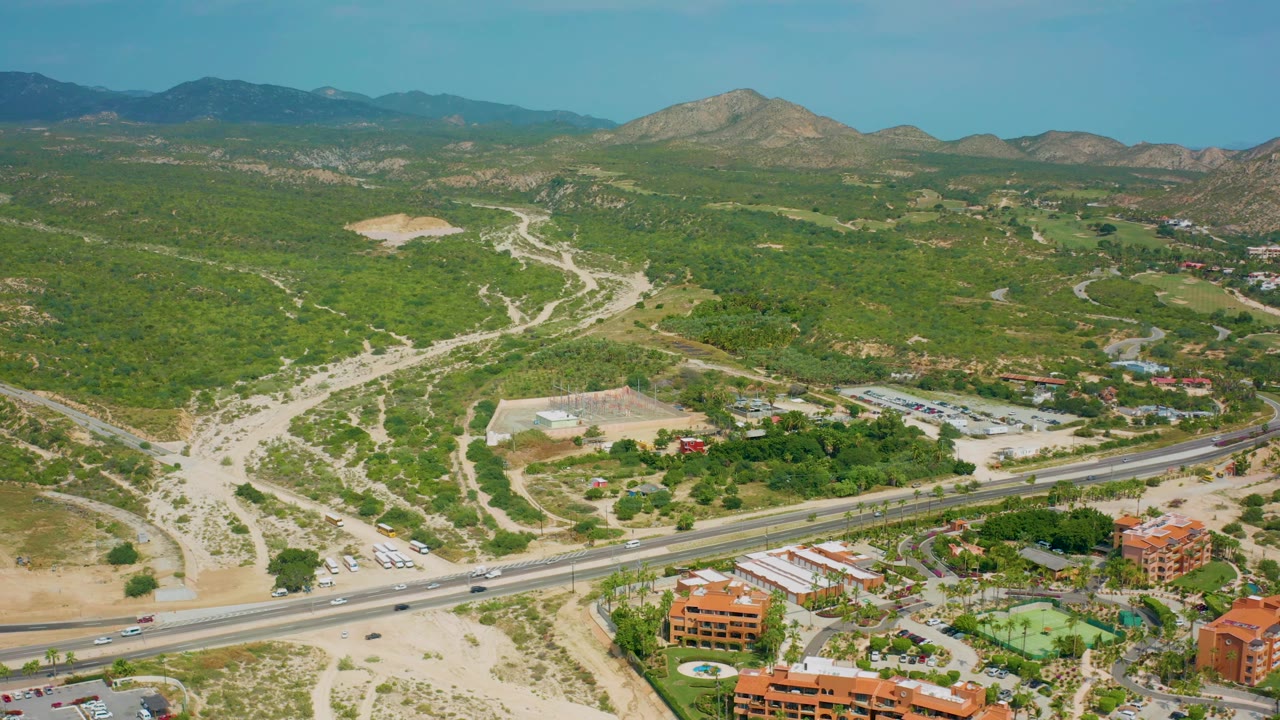Mid-clip aerial view of Desert Park Natural Reserve with small vehicles on the foreground highway, branching dirt tracks in pale soil, green vegetation, distant mountains, and no visible people.
