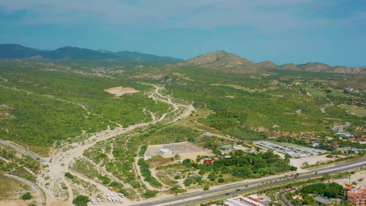 Second aerial frame of Desert Park Natural Reserve in Baja California Sur, Mexico, centering branching pale dirt roads across green desert hills, with the highway and parking lot still visible below and no visible people.