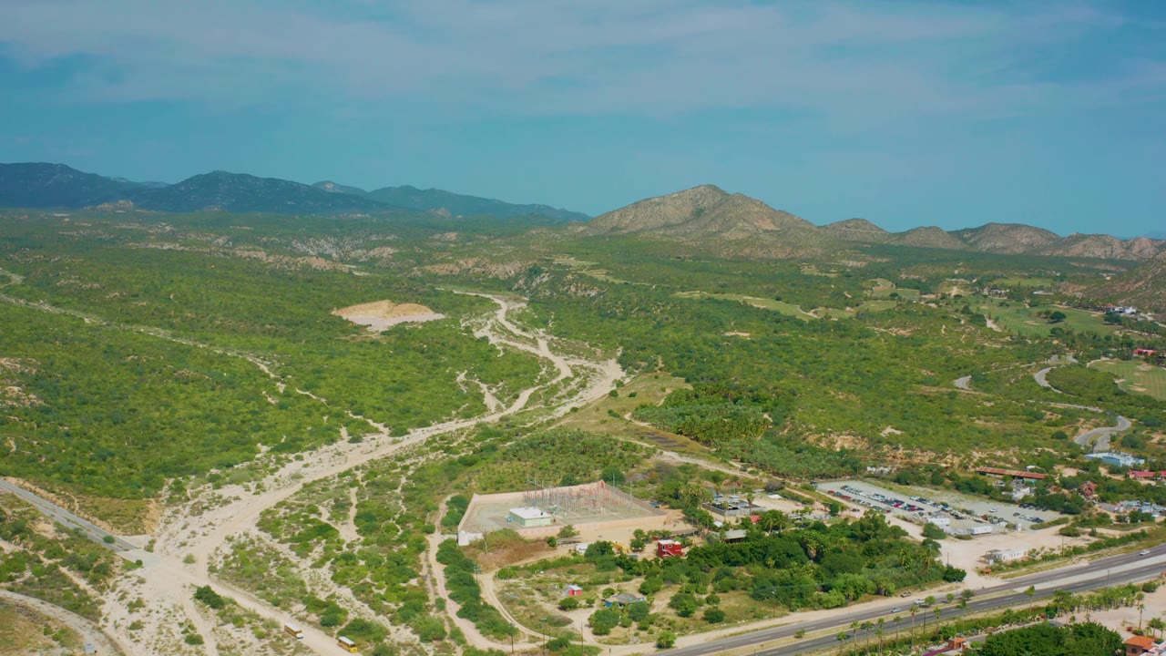 Fourth aerial frame of Desert Park Natural Reserve in Baja California Sur, Mexico, showing sunlit green hills, a network of dirt tracks, a foreground highway, a parking lot, scattered buildings, and no visible people.