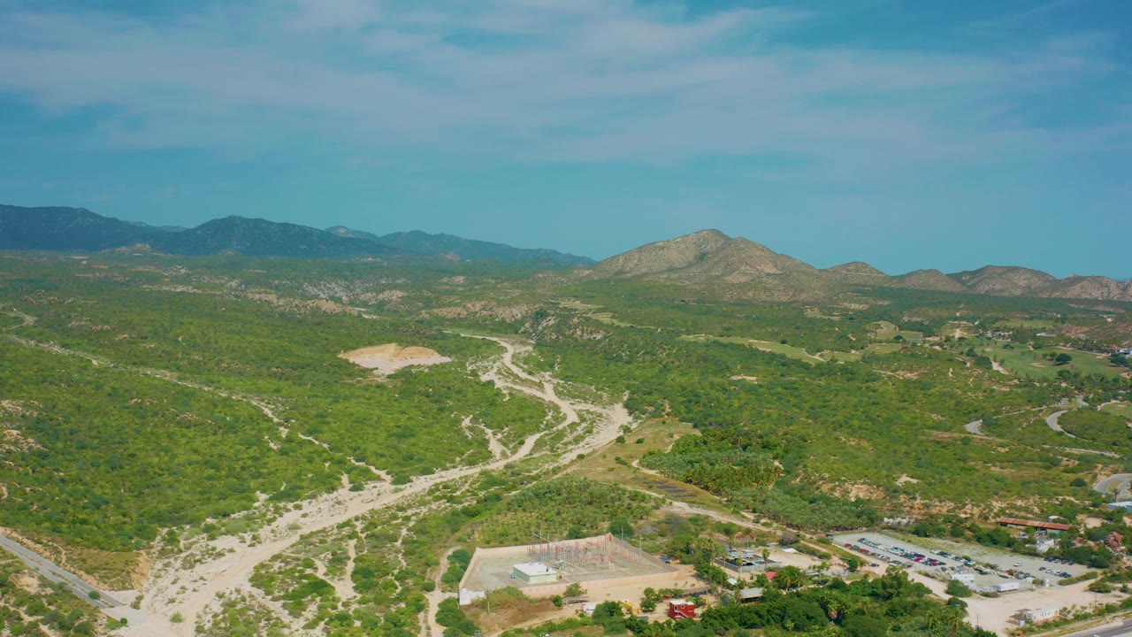 Opening aerial frame over Desert Park Natural Reserve in Baja California Sur, Mexico, showing pale dirt roads crossing green hills, distant mountains, blue sky, and no visible people.