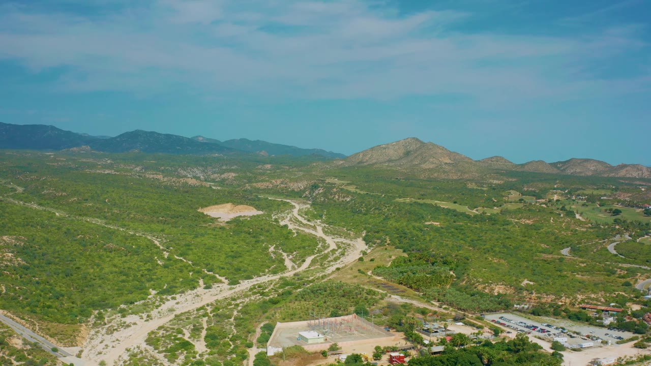 Wide aerial frame over Baja California Sur terrain with a small developed area near the lower edge, winding roads through green hills, distant mountains, and no visible people.