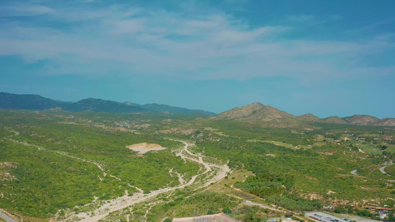 Aerial view one second into the clip, with a broad sweep of green desert hills, pale road forks, mountain ridges on the horizon, and no visible people.