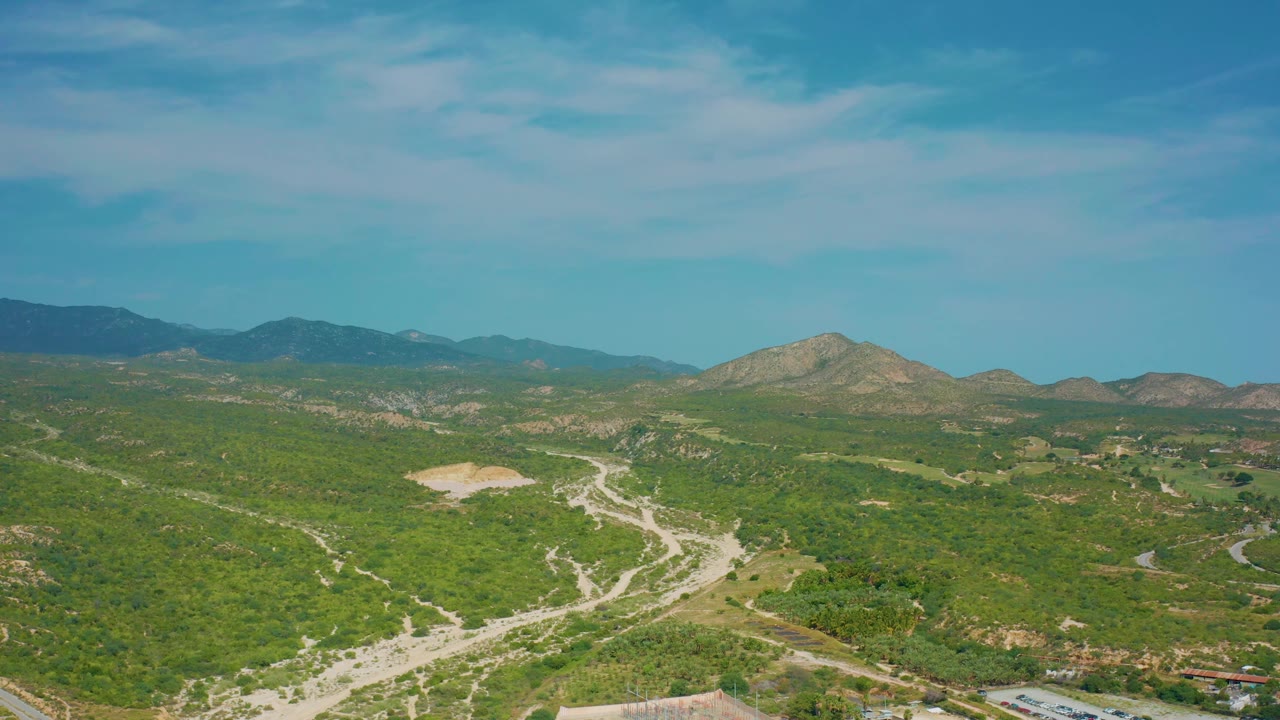 Aerial frame over Baja California Sur terrain showing a light-colored dirt clearing, branching road cuts across green hills, layered mountains, and no visible people.