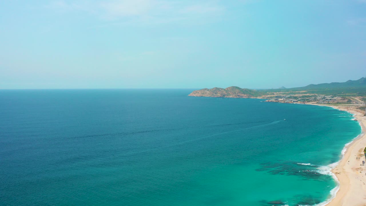 Opening aerial frame over Desert Park Natural Reserve in Baja California Sur, with deep blue ocean, turquoise shallows, a curving sandy beach, distant arid headland, and no visible people.