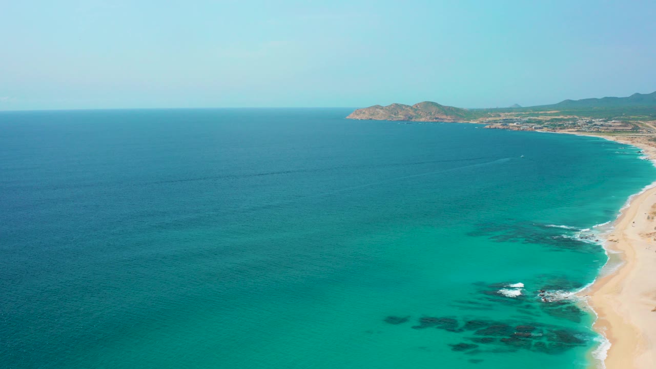 Aerial view over Desert Park Natural Reserve in Baja California Sur, with turquoise shallows, deeper blue ocean, a sandy beach on the right, a distant headland, and no visible people.