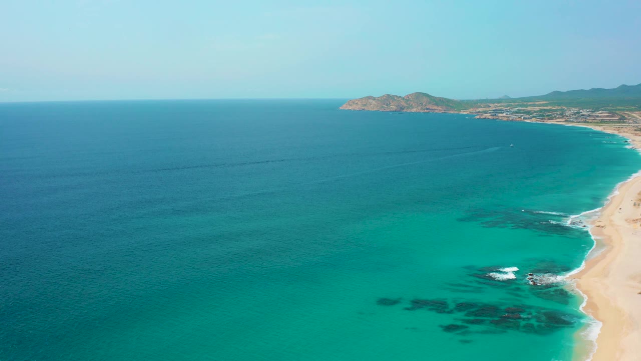 High aerial frame from Desert Park Natural Reserve in Baja California Sur, showing turquoise coastal water, white surf at the sand line, distant hills and headland, and no visible people.