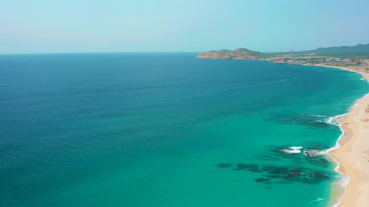 Wide aerial view at Desert Park Natural Reserve in Baja California Sur, with an empty-looking sandy beach edge, teal water, darker submerged areas, a rocky headland, and no visible people.