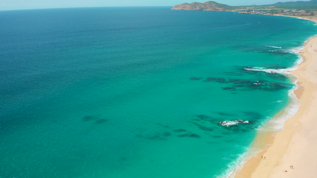 High aerial view over Desert Park Natural Reserve in Baja California Sur, with turquoise water, dark submerged rocks, pale sand, distant shoreline, and no visible people at this moment.