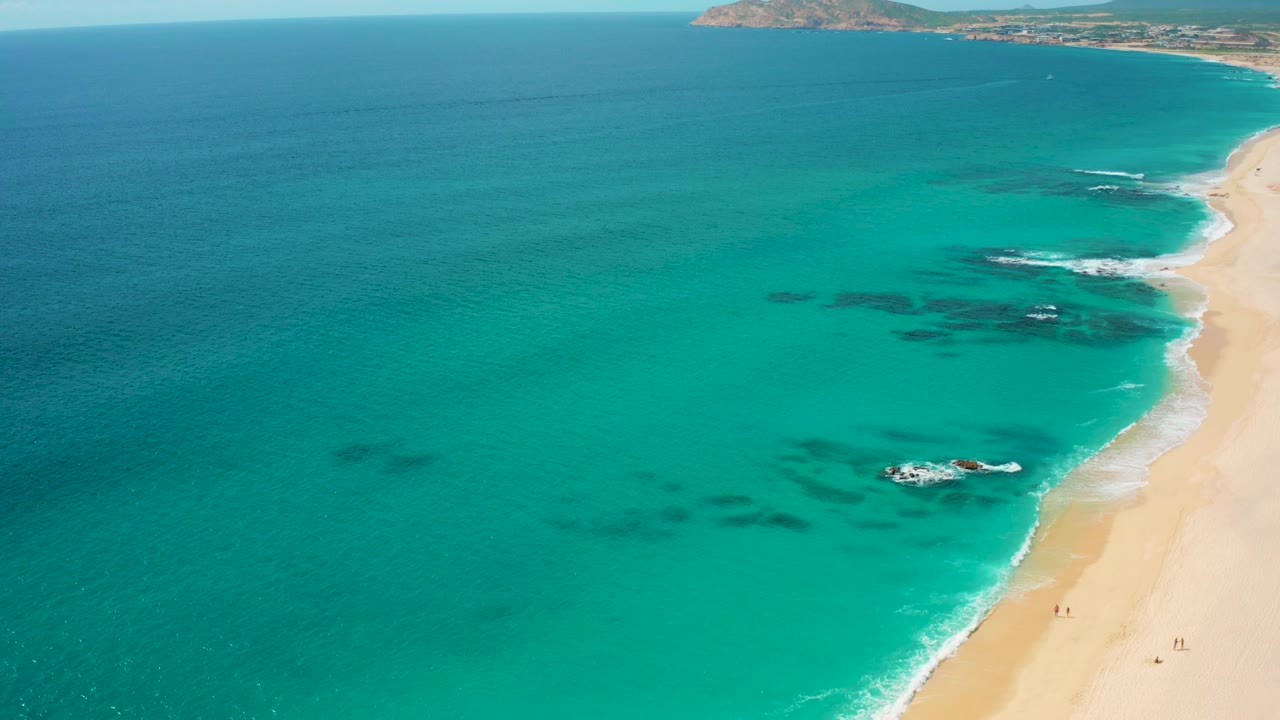 High drone frame over Desert Park Natural Reserve in Baja California Sur with broad turquoise water, dark submerged formations, a bright sandy beach along the right, white wave edges, and small walkers on the sand.
