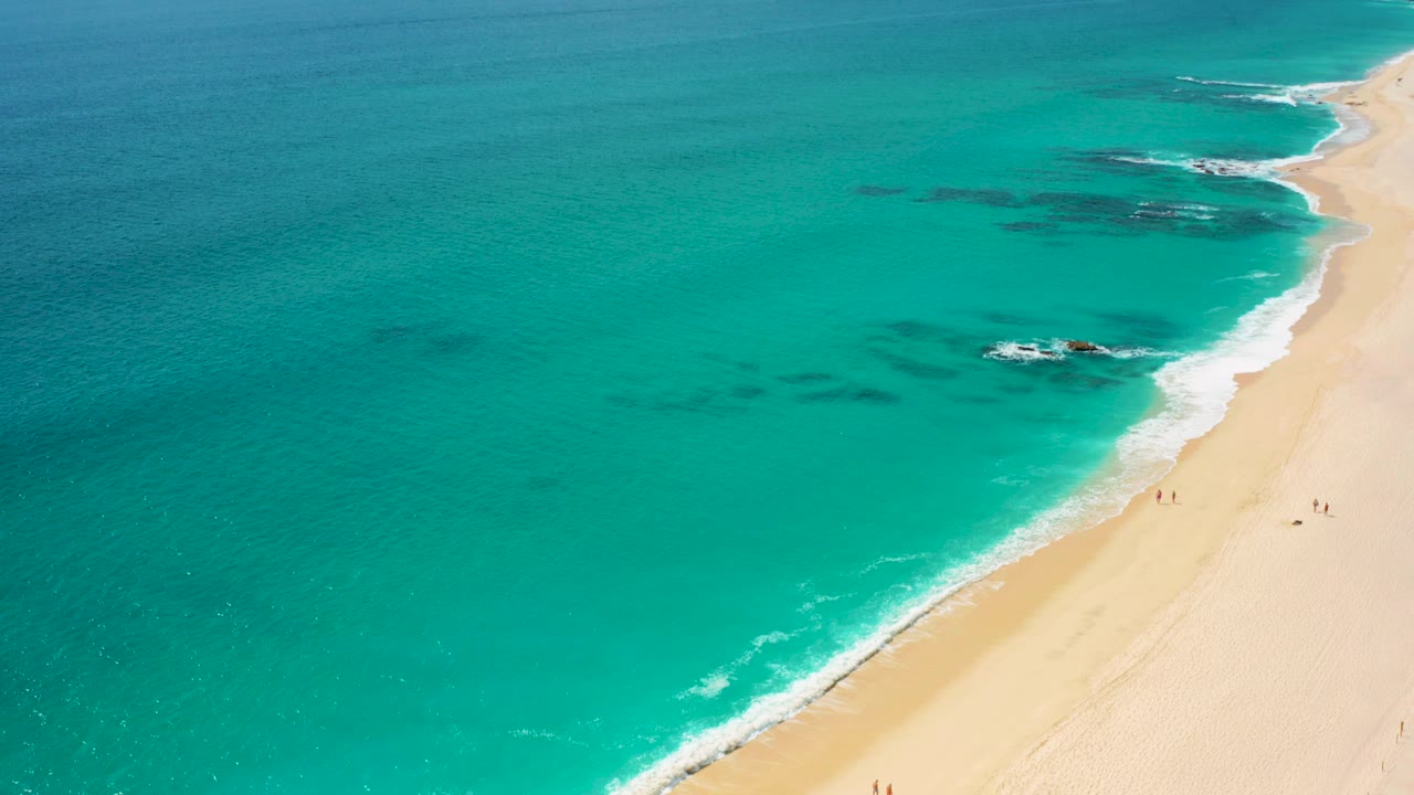 Opening aerial frame over Desert Park Natural Reserve in Baja California Sur, Mexico, with turquoise ocean on the left, pale sand on the right, offshore rock patches, and tiny beach walkers near the shore.