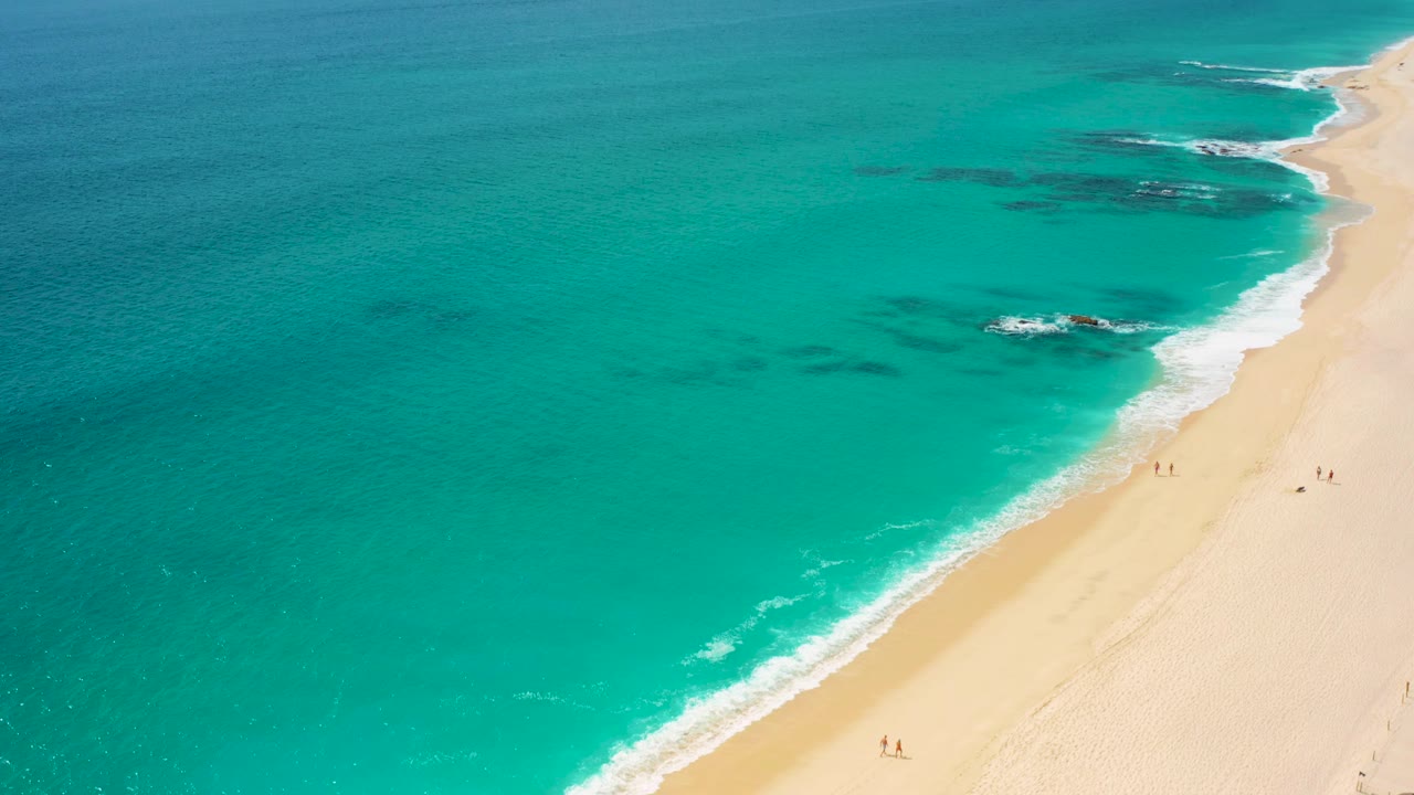 High-angle frame at Desert Park Natural Reserve with clear turquoise water, pale beach on the right, dark submerged patches offshore, foamy surf, and scattered tiny figures walking along the sand.
