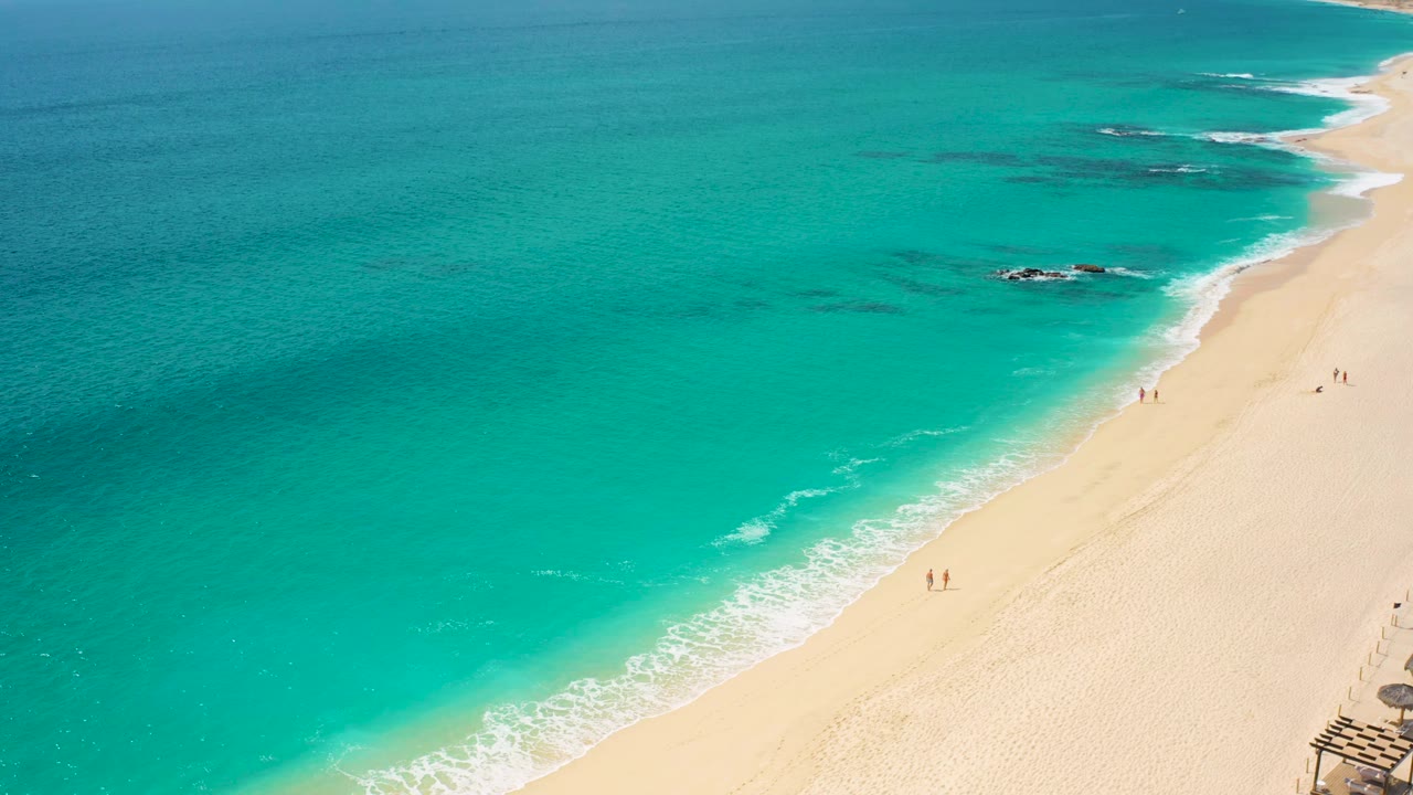 Aerial coastline with turquoise water, pale beach, and small figures on sand.