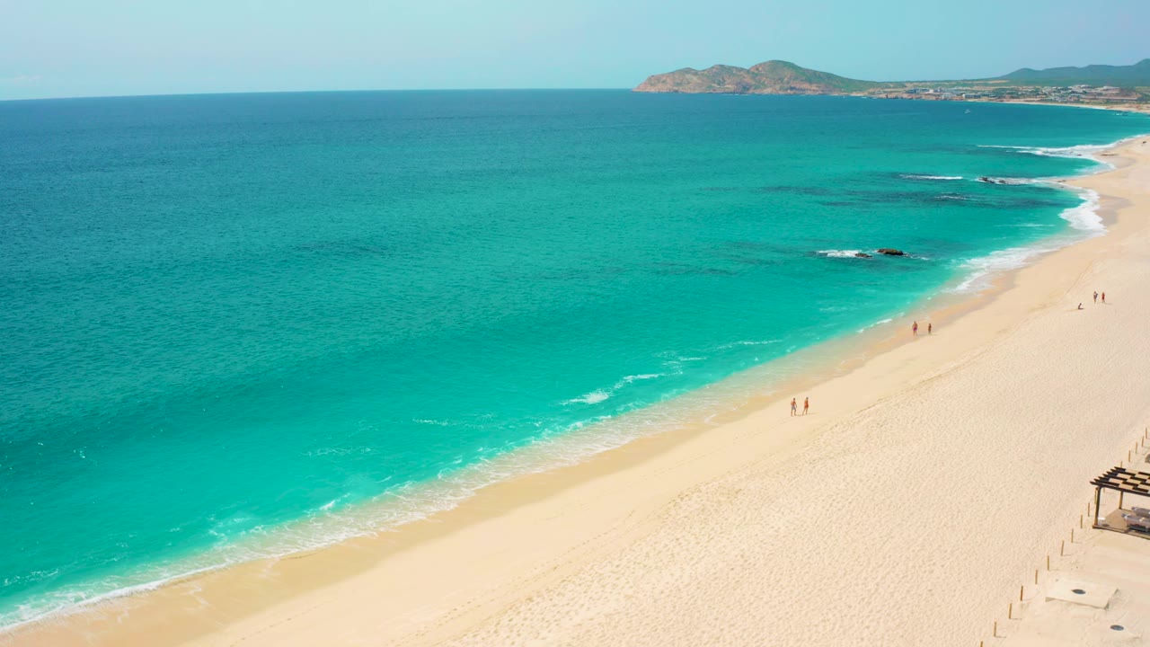 Aerial view in Baja California Sur with the shoreline running diagonally across the frame, white wave edges on the sand, and small distant walkers along the beach.