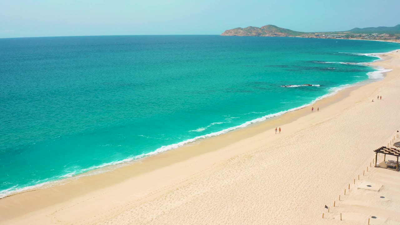 Baja California Sur coast from above, with broad turquoise water on the left, a clean sandy beach on the right, and a low rocky headland across the upper background.