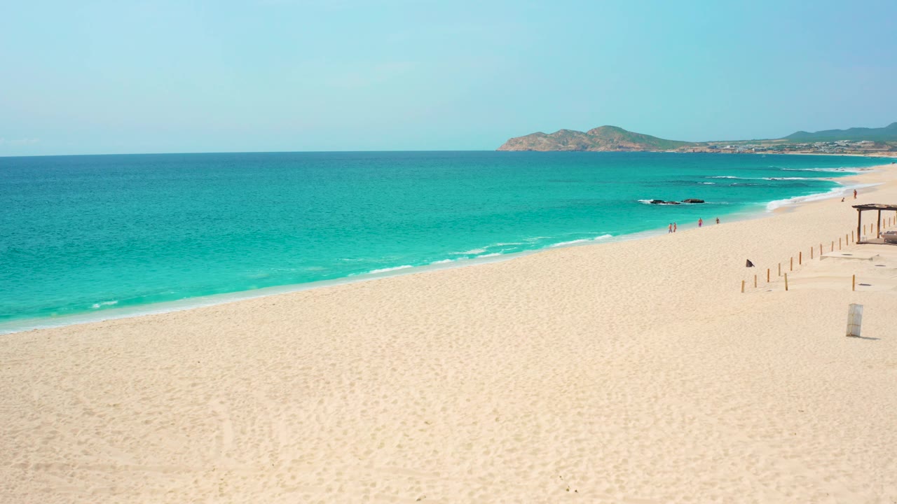 Wide Baja California Sur beach frame with open sand in the foreground, bright turquoise ocean across the middle, a low headland on the horizon, right-side posts, and no visible people.