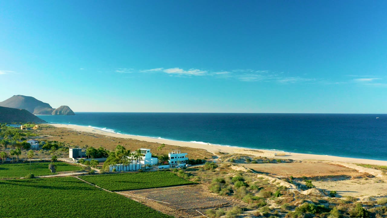 Opening aerial frame over Todos Santos BCS Mexico showing green fields and white buildings below, a long sandy beach, blue ocean, distant mountains, and no visible people.
