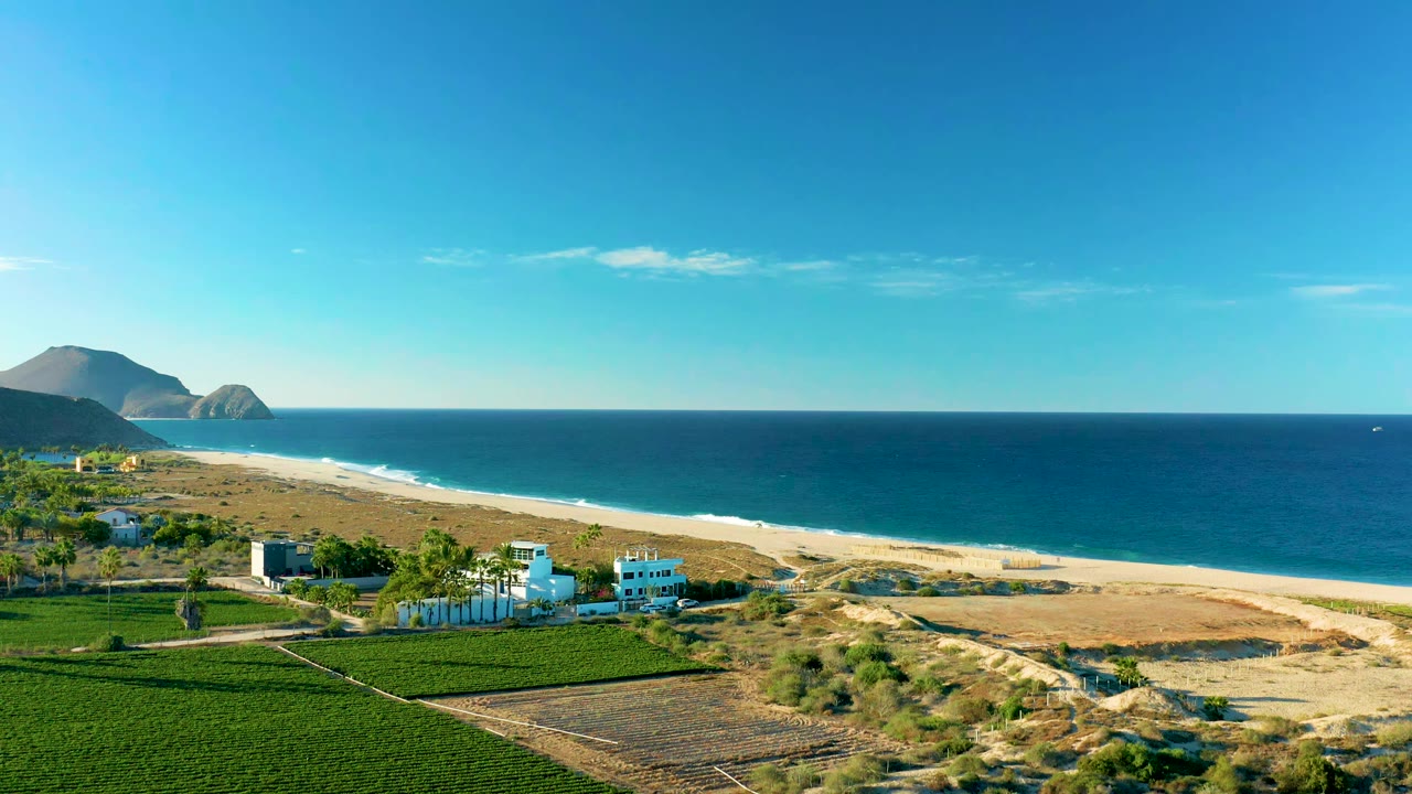 Aerial view in Todos Santos BCS Mexico with white buildings and green fields lower left, dry dunes beside the beach, blue ocean across the right, mountains in the distance, and no visible people.