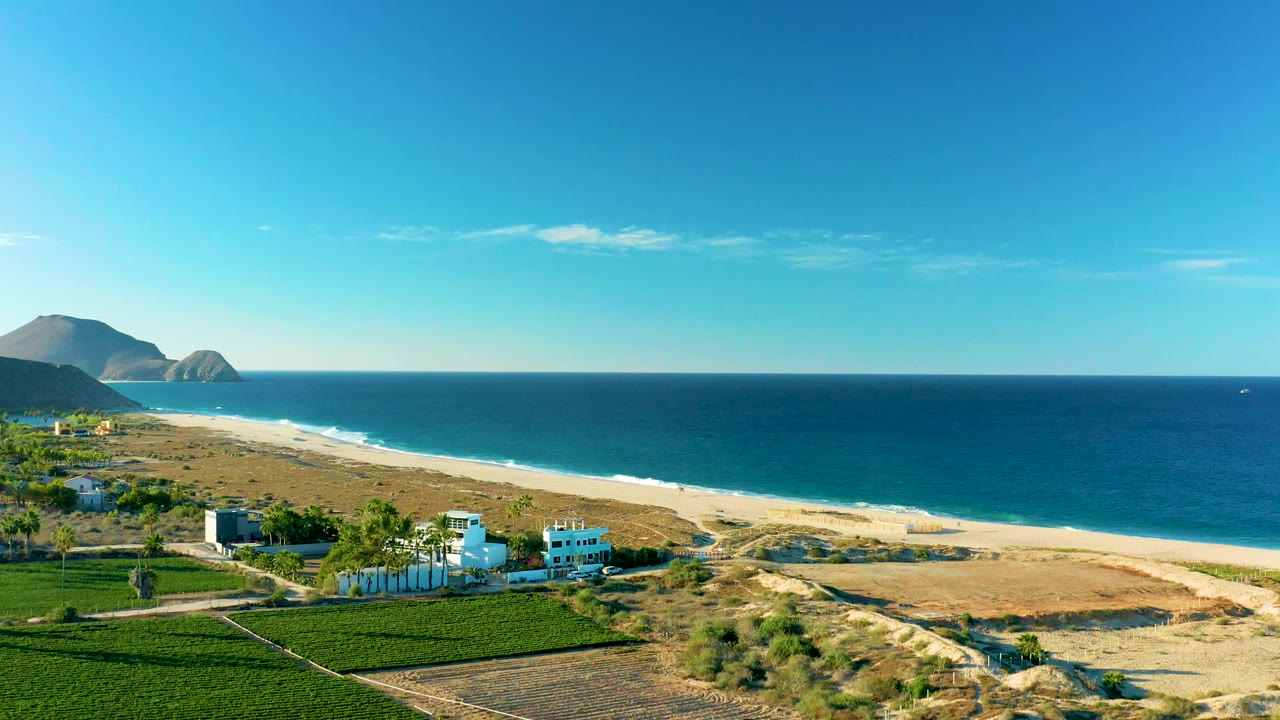 Todos Santos BCS Mexico aerial frame with the buildings shifted lower in the composition, green fields at left, pale sand and dunes through the center, deep blue ocean, and no visible people.