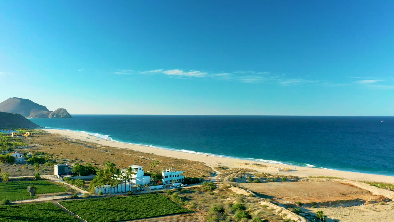 Wide aerial coastline with ocean, surf line, dry dunes, and buildings at lower left.