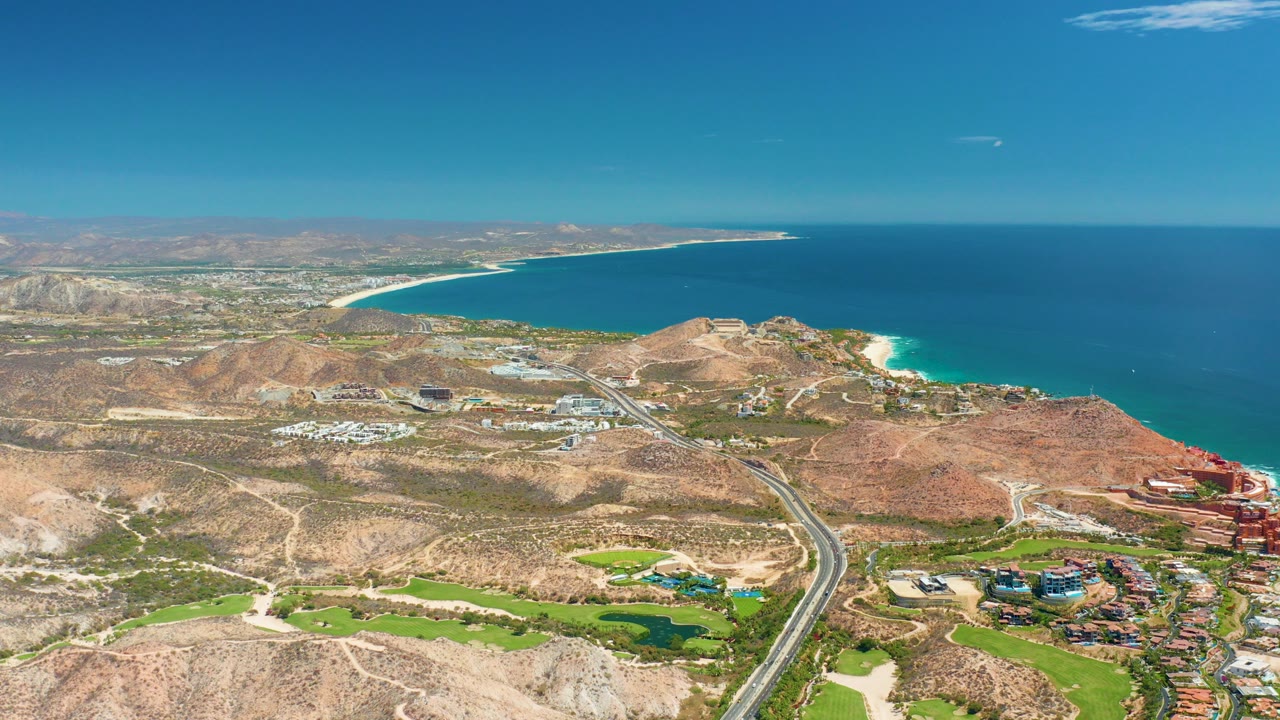 Wide aerial view over Marquis Los Cabos BCS Mexico, with dry hills in front, a road leading through developed green grounds, blue ocean beyond, and no visible people.