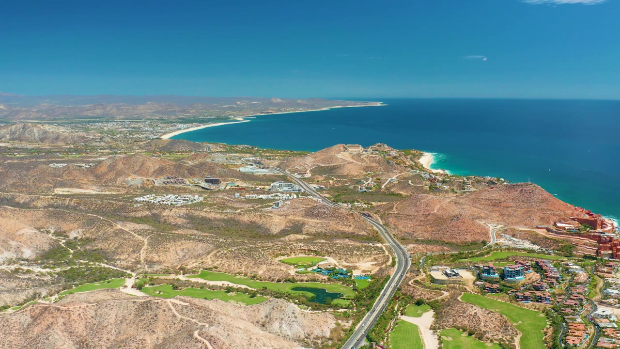 Aerial frame over Marquis Los Cabos BCS Mexico with the paved road cutting between arid hills and green grounds, turquoise surf along the coast, and no visible people.