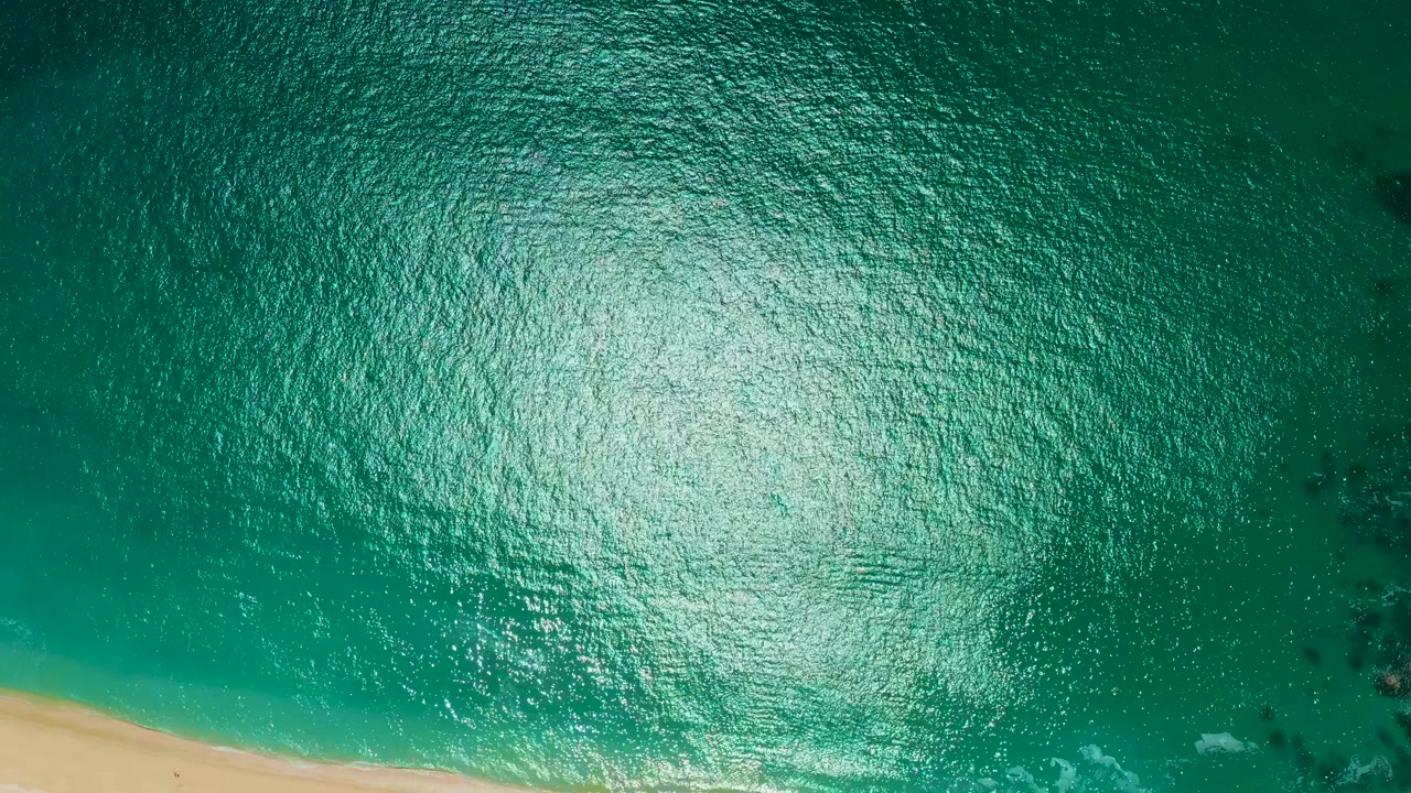 Overhead teal water fills the frame with sun glare across the surface and the sandy shoreline still visible along the lower left, with no visible people.