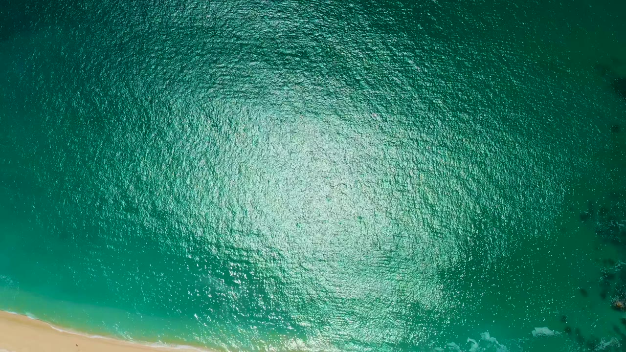 A top-down frame shows bright turquoise water, a pale sand strip near the bottom left, faint wave foam, and no visible people at Marquis Los Cabos BCS Mexico.