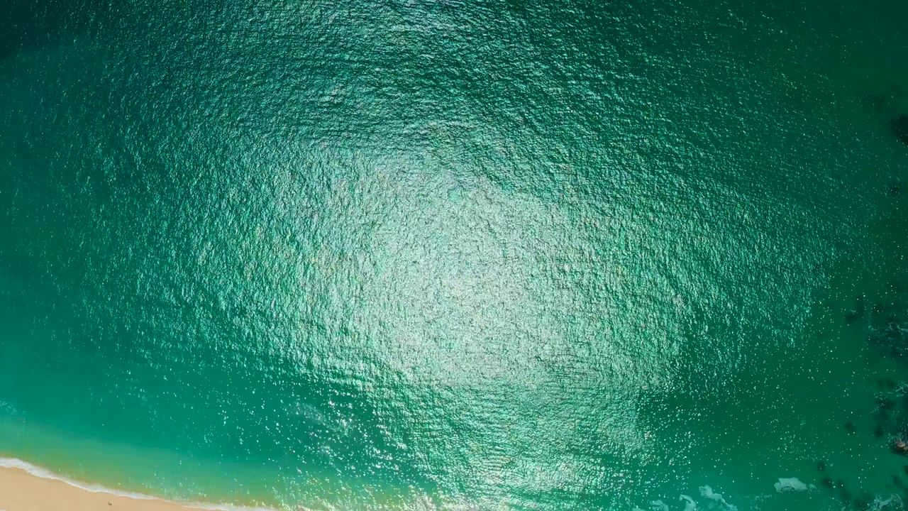 Sun reflection covers the rippled ocean surface while the sand line appears in the lower left corner and darker patches sit under the water, with no visible people.