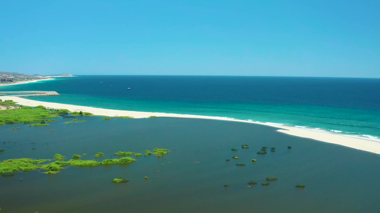 Aerial frame from San Jose del Cabo Estuary, BCS Mexico, showing more of the lagoon foreground, scattered green plant islands, a long white beach, blue-green surf bands, open ocean, and no visible people.