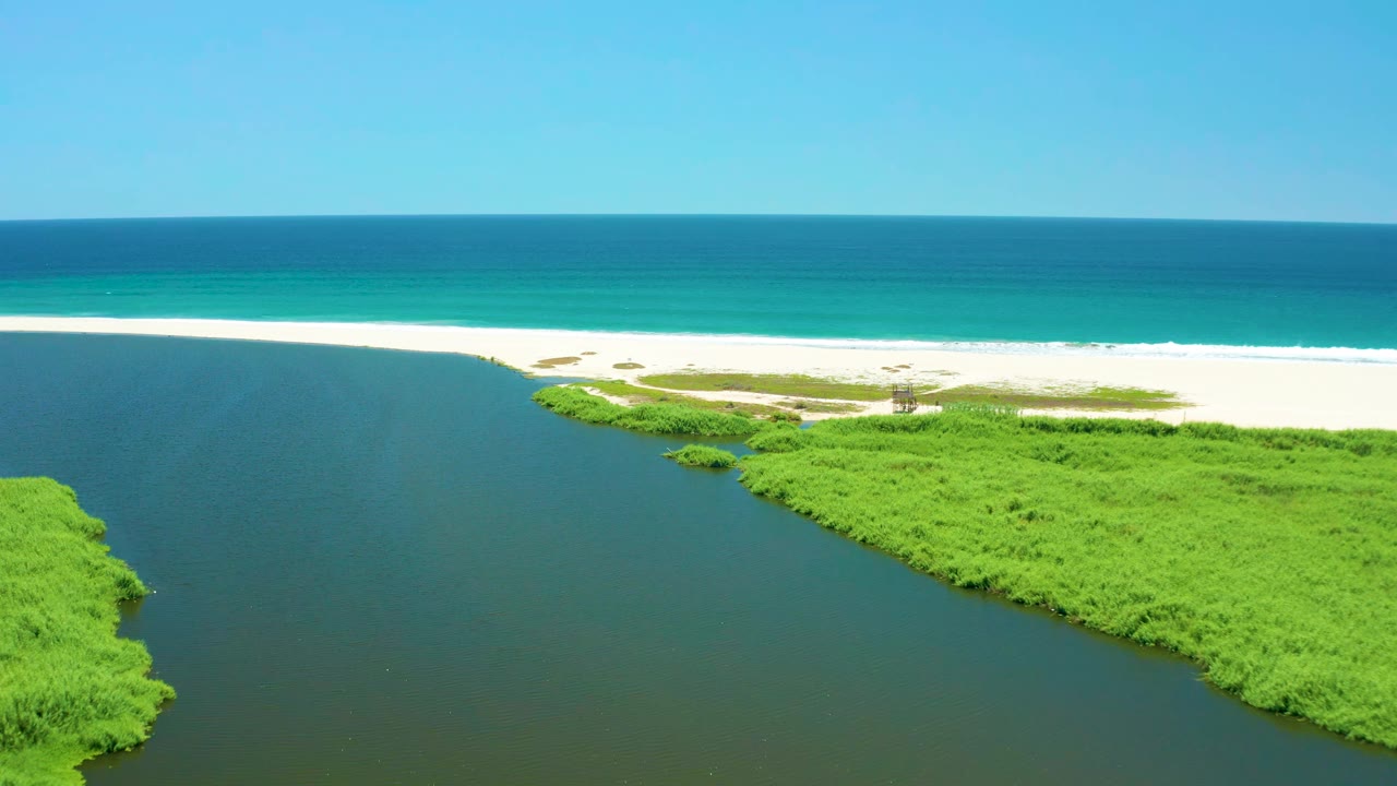 Aerial view of the San Jose Del Cabo Estuary one second in, with the inlet channel opening toward a white sandbar, bright green mangroves, turquoise water, and no visible people.