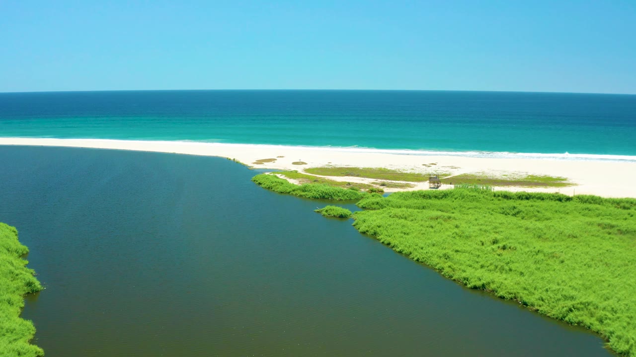 Aerial frame of the San Jose Del Cabo Estuary with the shoreline shifted across the lower right, open inlet water on the left, pale sand in the center, turquoise ocean, and no visible people.