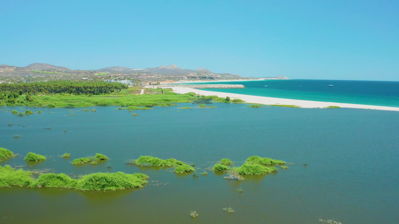 Wide aerial frame of the San Jose del Cabo Estuary in Mexico, showing small green vegetation islands in lagoon water, a long sandbar-like beach, ocean water, hills, and no visible people.