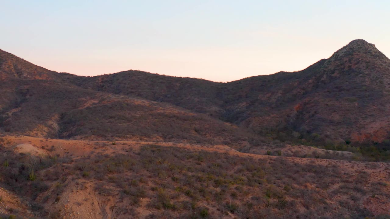 Wide Cabo Pulmo BCS Mexico landscape showing reddish-brown desert hills, scattered low plants, shadowed ridges, a pale pink-blue sky, and no visible people.