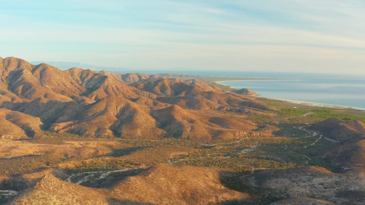 Aerial frame over Cabo Pulmo BCS Mexico showing brown ridges, scrubby valley terrain, distant coastal water, pale sky, and no visible people.
