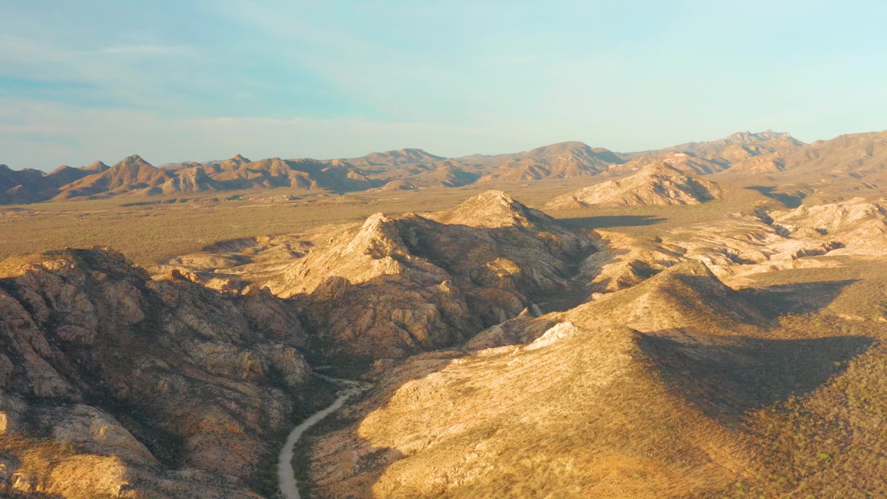 Opening aerial frame over Cabo Pulmo BCS Mexico shows rocky golden desert ridges, a pale winding road in the lower valley, distant mountains, clear sky, and no visible people.