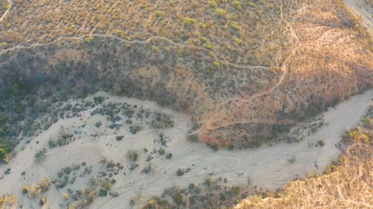 A Cabo Pulmo, Mexico aerial view shows the dry riverbed bending along a shadowed hillside with scattered shrubs and pale sand, with no visible people.