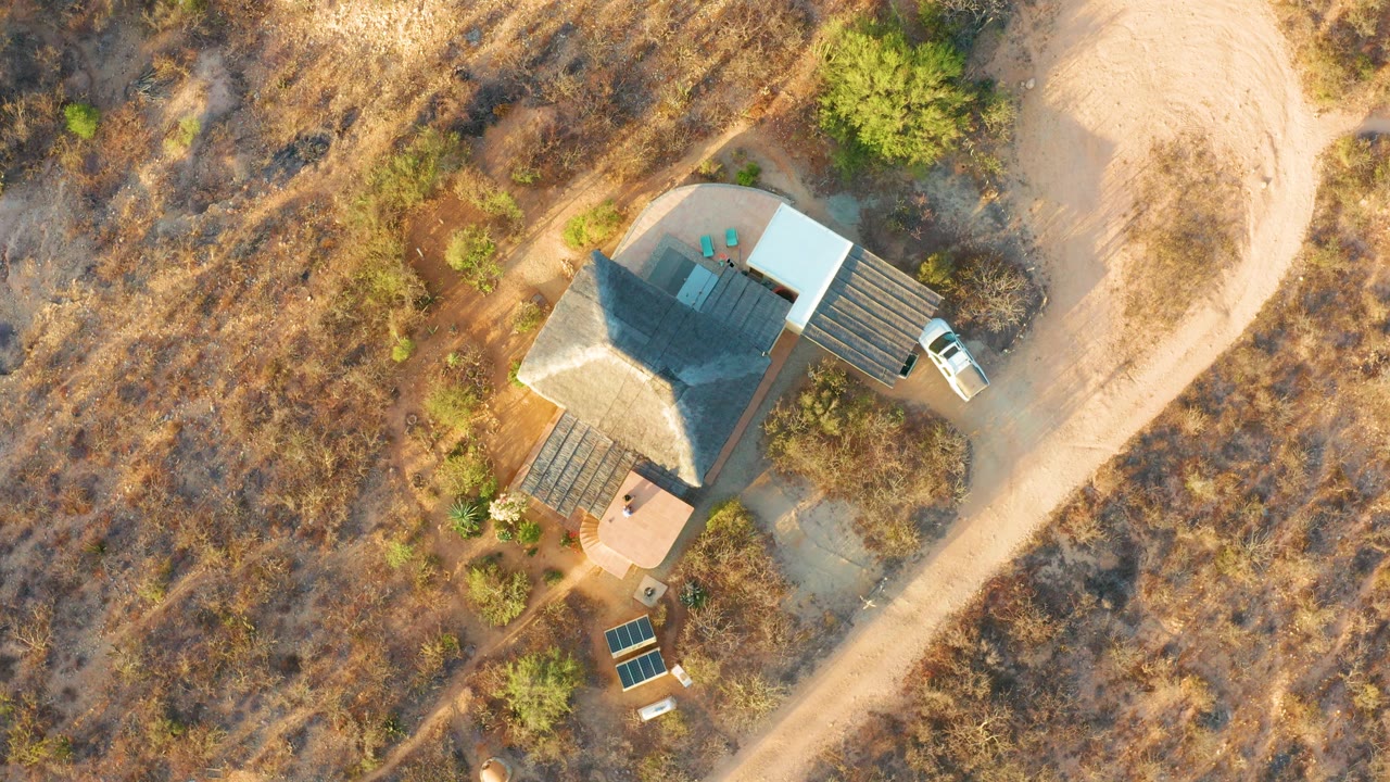 Overhead frame from Cabo Pulmo BCS Mexico showing a rural house with a dark roof, white roof section, nearby deck structures, a white truck by the driveway, dry brown grassland, and no visible people.