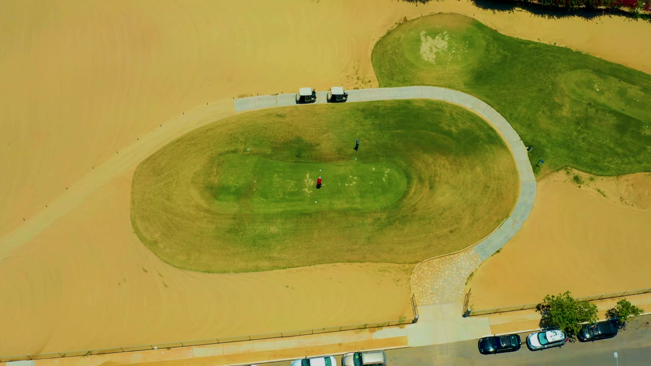 Aerial view of two people on a small grass oval surrounded by sand and a road edge.