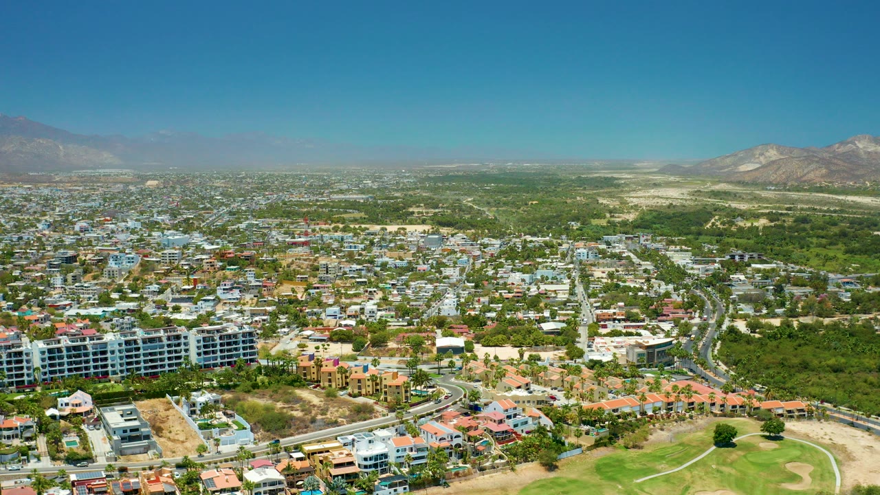 Opening aerial frame over San Jose del Cabo BCS Mexico with low-rise buildings, curving roads, green foreground, hazy mountains, clear blue sky, and no visible people.