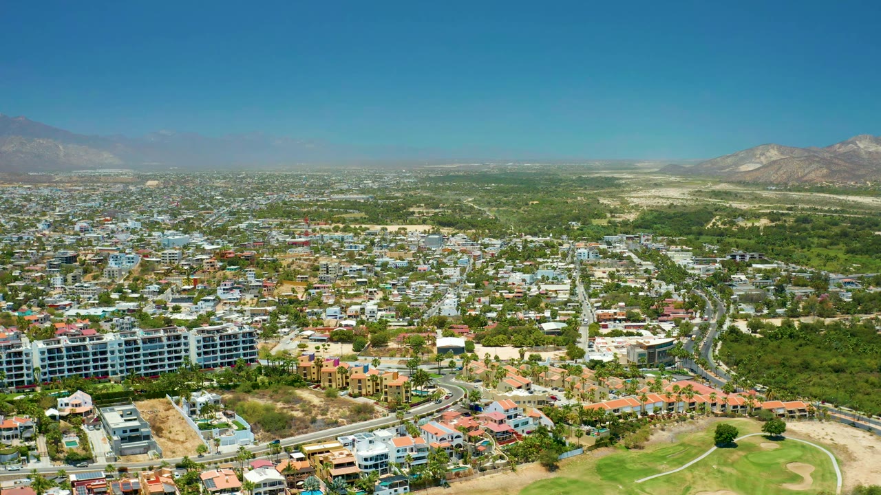 One second into the San Jose del Cabo BCS Mexico aerial, the view holds over clustered buildings, a central road corridor, green open space, distant mountains, and no visible people.