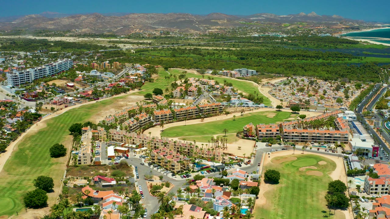 High aerial frame over San Jose del Cabo BCS Mexico showing golf fairways, sand traps, red-roofed residences, dense coastal greenery, distant ocean, hills, and no visible people.