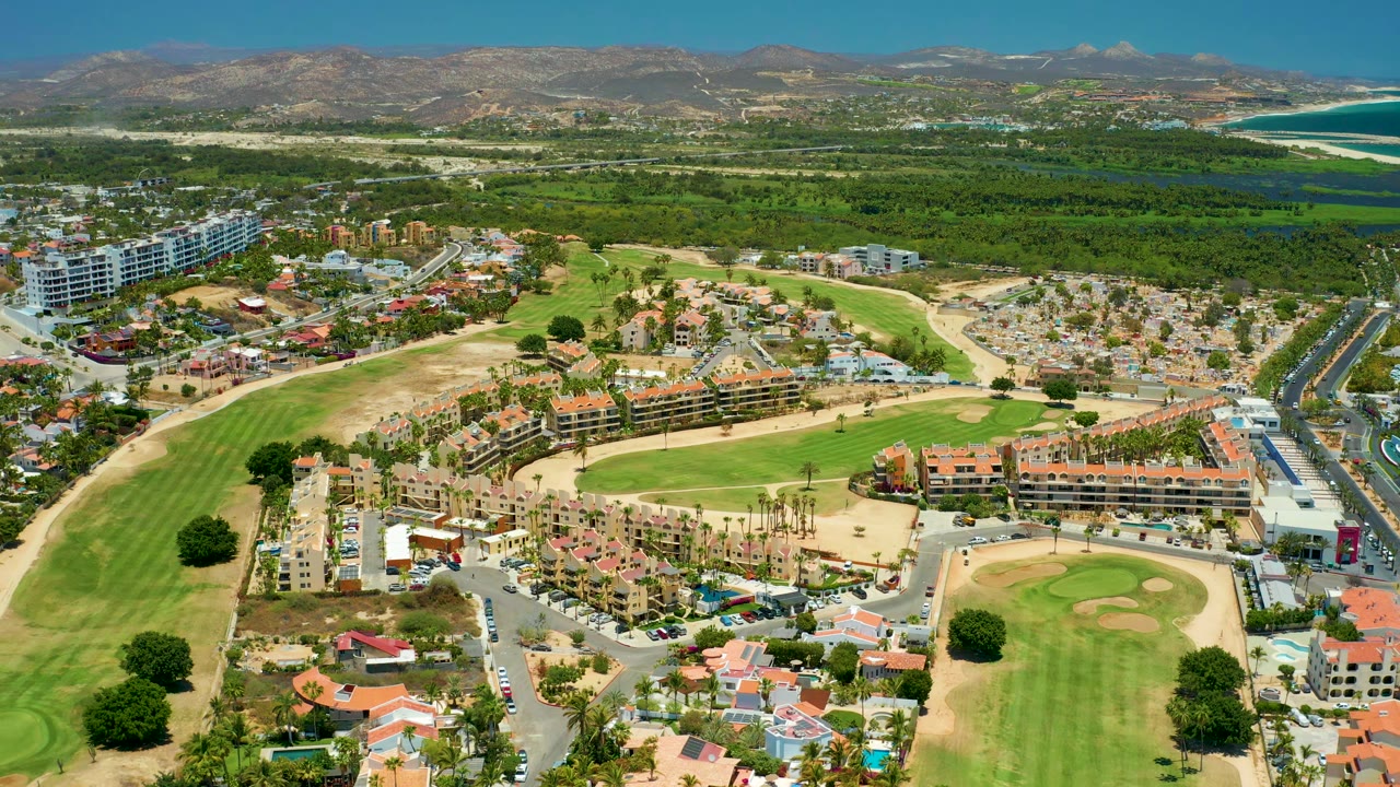 Drone view across golf course, apartment blocks, roads, beach edge, and hills.