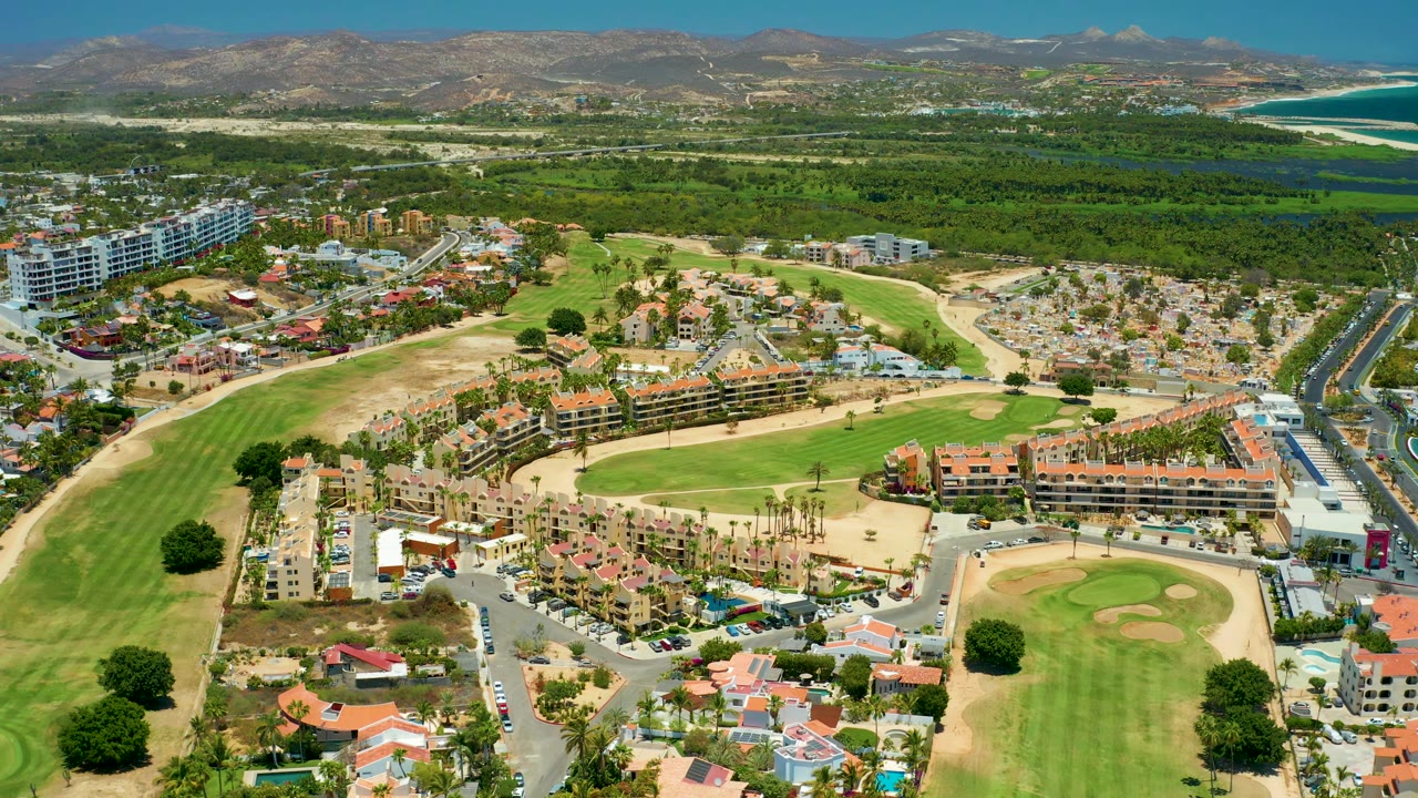 San Jose del Cabo BCS Mexico aerial frame with golf greens and bunkers below, clustered residences and parking areas in the middle, coastal vegetation and blue ocean beyond, and no visible people.