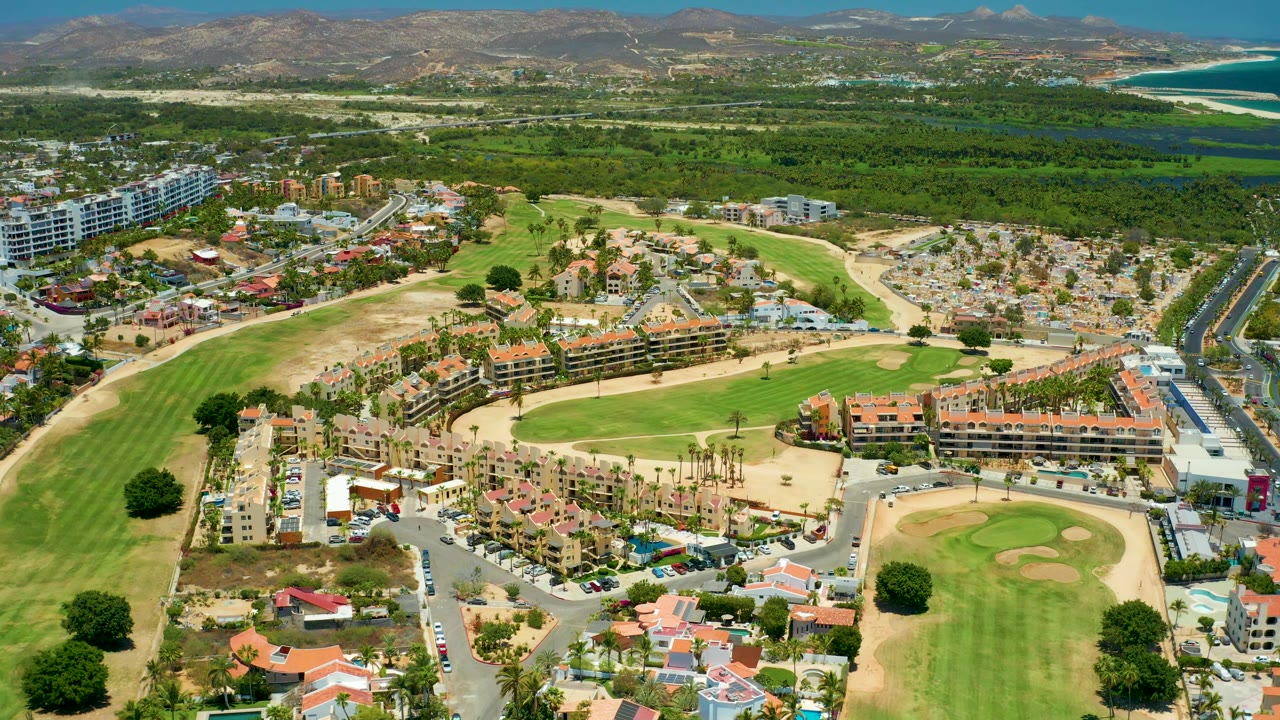 Aerial frame over golf course housing, roads, coastal greenery, ocean, and hills.