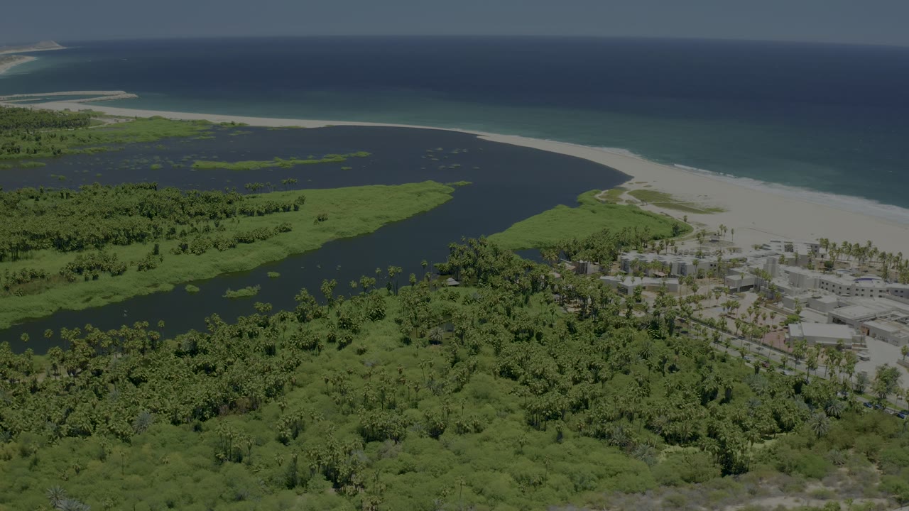 Mid-clip aerial frame over San Jose del Cabo, Mexico with broad lagoon water, mangroves below, a sandy beach band, blue ocean, and no visible people.