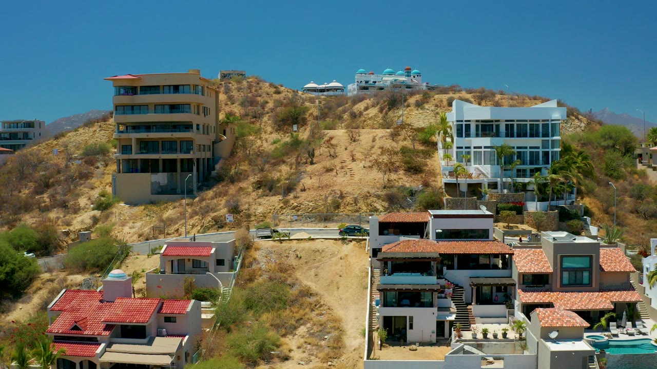 Opening frame of San Jose del Cabo, Mexico hillside homes with red tile roofs, tan and white buildings, dry scrub, a pale winding road, and no visible people.