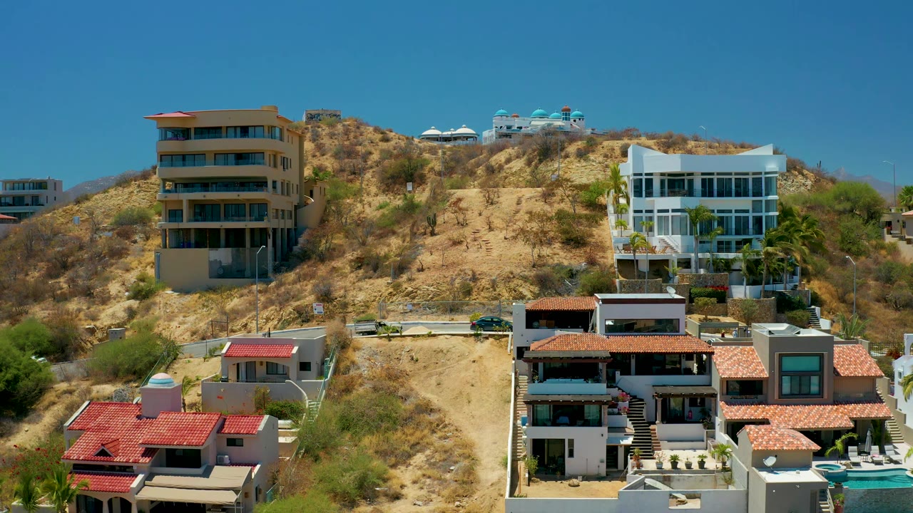 The aerial view holds on San Jose del Cabo hillside houses, with a white glass-front building at right, a tan building at left, dry terrain, and no visible people.
